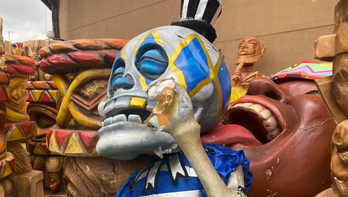 Grote carnavalsfiguur van een blauw-grijze schedel met felgekleurde details, zittend tussen andere beschadigde decorstukken.