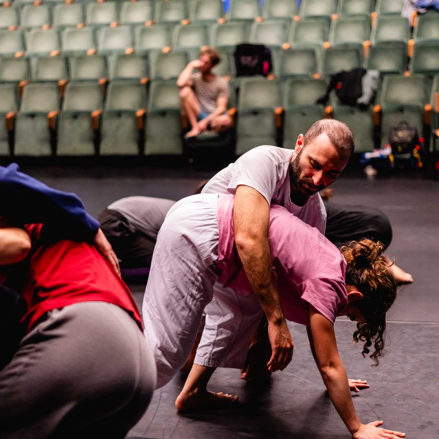 Dansers werken samen in een fysieke workshop op het podium van een theaterzaal, met lege tribunes op de achtergrond.