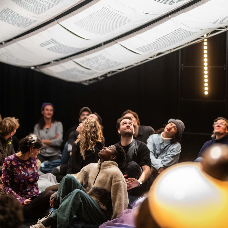 Foto Welcome To Our Guesthouse #8 - Een groep bezoekers zit dicht bij elkaar en kijkt omhoog naar teksten die boven hen hangen, in een intieme en aandachtige setting.