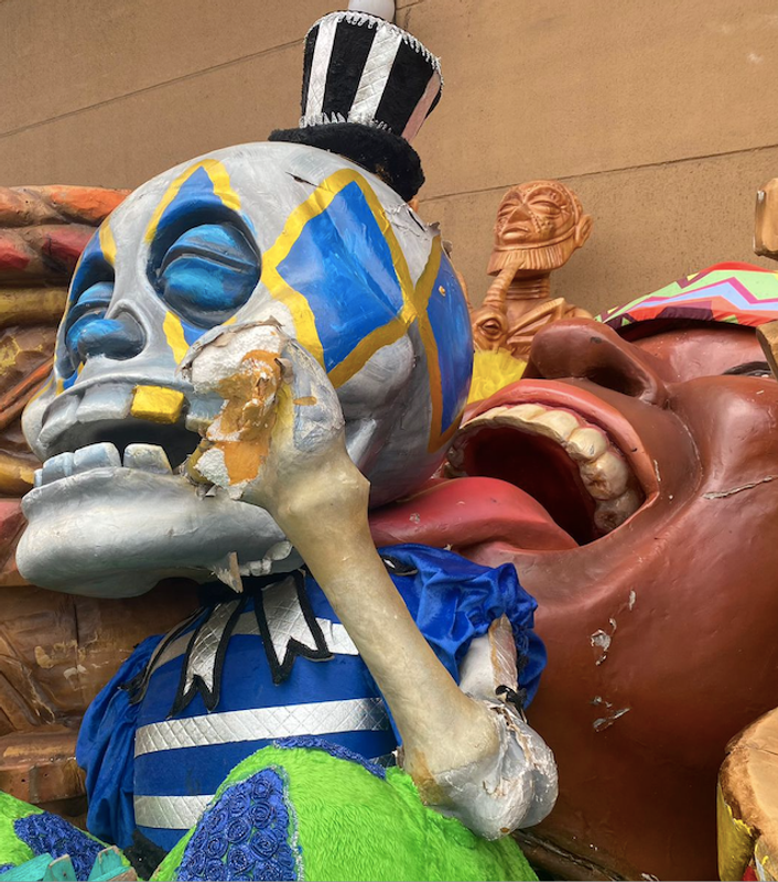 Grote carnavalsfiguur van een blauw-grijze schedel met felgekleurde details, zittend tussen andere beschadigde decorstukken.