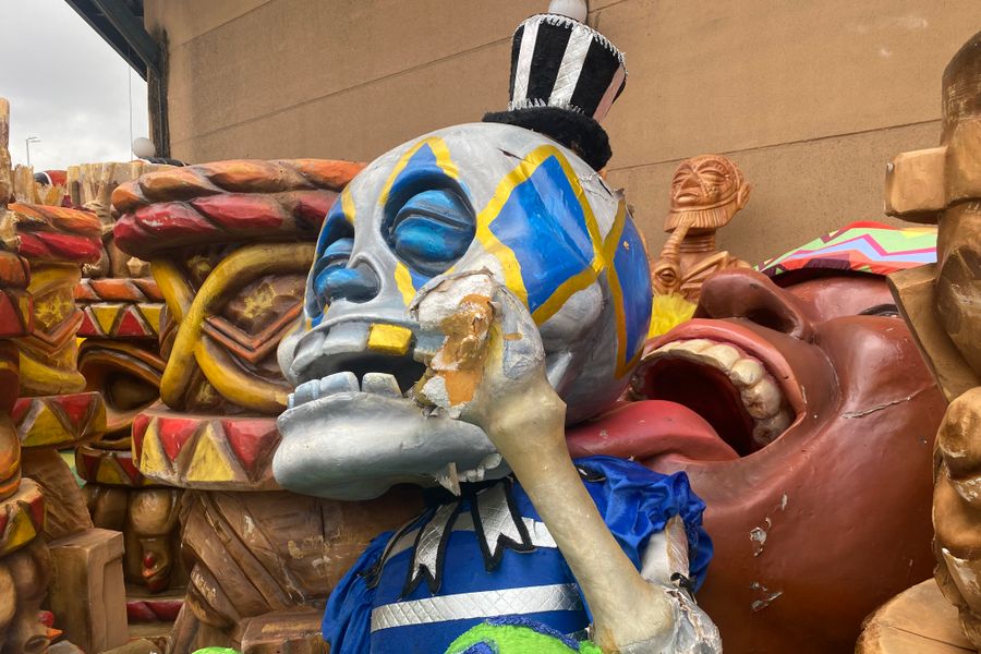 Grote carnavalsfiguur van een blauw-grijze schedel met felgekleurde details, zittend tussen andere beschadigde decorstukken.