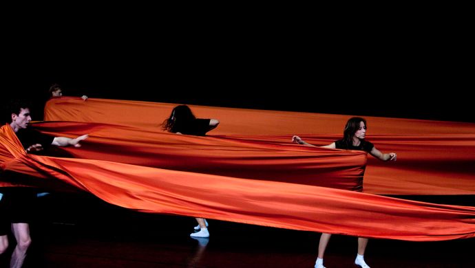 Vier performers op een donker podium trekken en dragen een lange, golvende strook oranje stof, die hen met elkaar verbindt en spanning en beweging zichtbaar maakt.