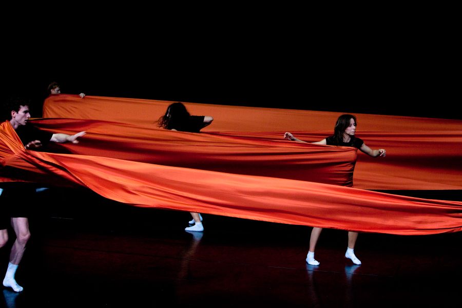 Vier performers op een donker podium trekken en dragen een lange, golvende strook oranje stof, die hen met elkaar verbindt en spanning en beweging zichtbaar maakt.