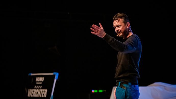 De Cock staat op een podium met een microfoonheadset en maakt een expressief gebaar met zijn hand, alsof hij het publiek toespreekt of een punt kracht bijzet. Op de achtergrond is een bord zichtbaar met "Rock Werchter".