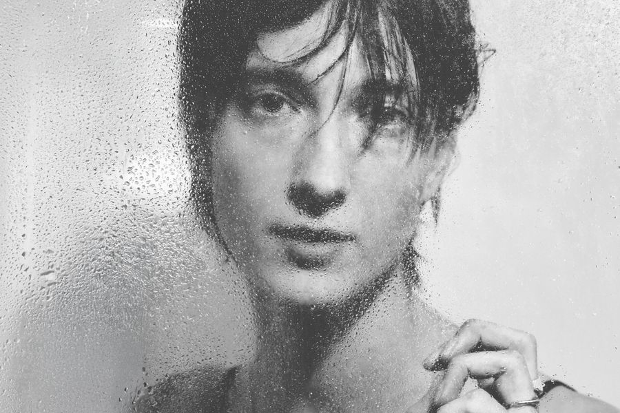 Zwart-wit portret van een persoon achter een beslagen en nat oppervlak, met waterdruppels op het glas en een dromerige, zachte blik.