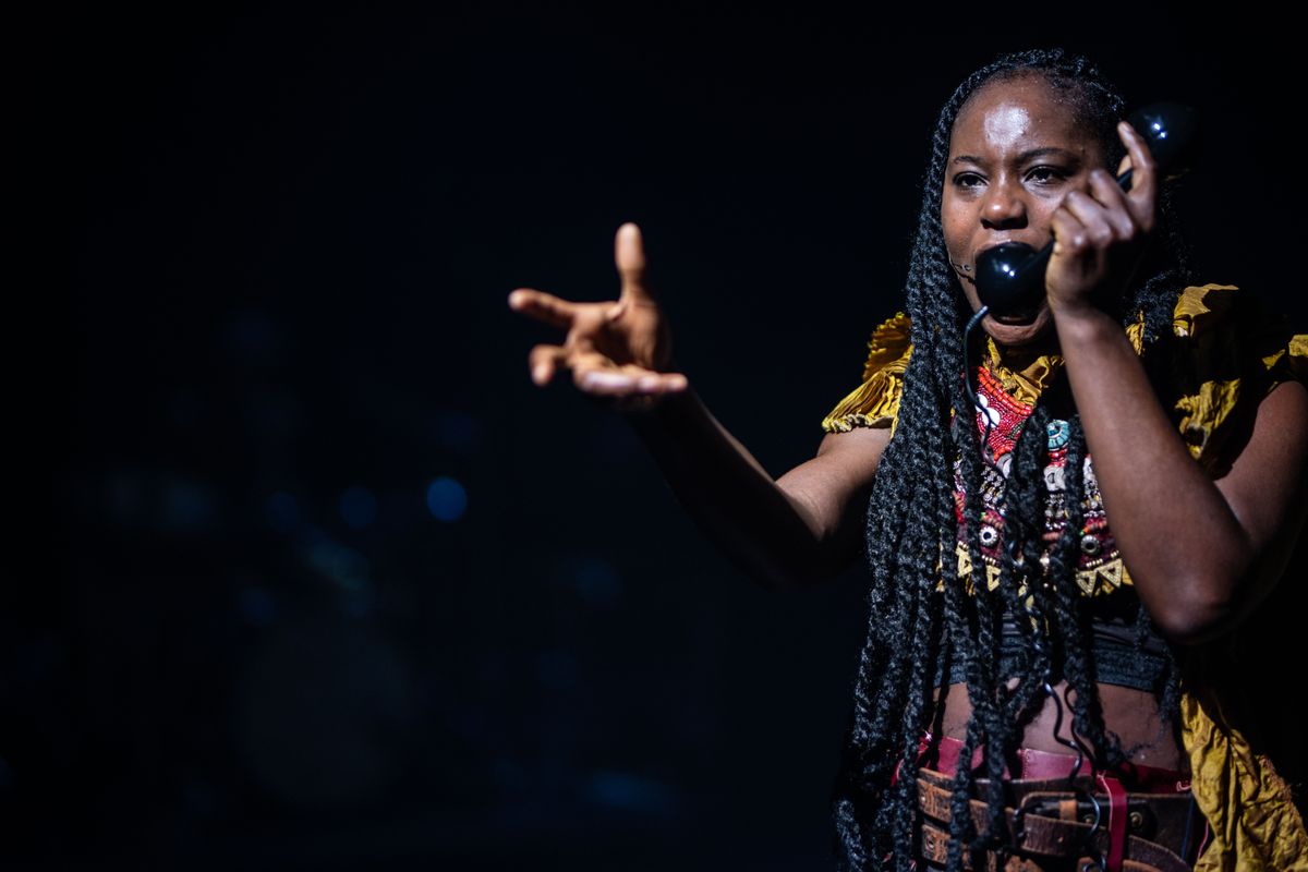 Een performer met lange vlechten spreekt intens terwijl zij een ouderwetse telefoonhoorn vasthoudt.