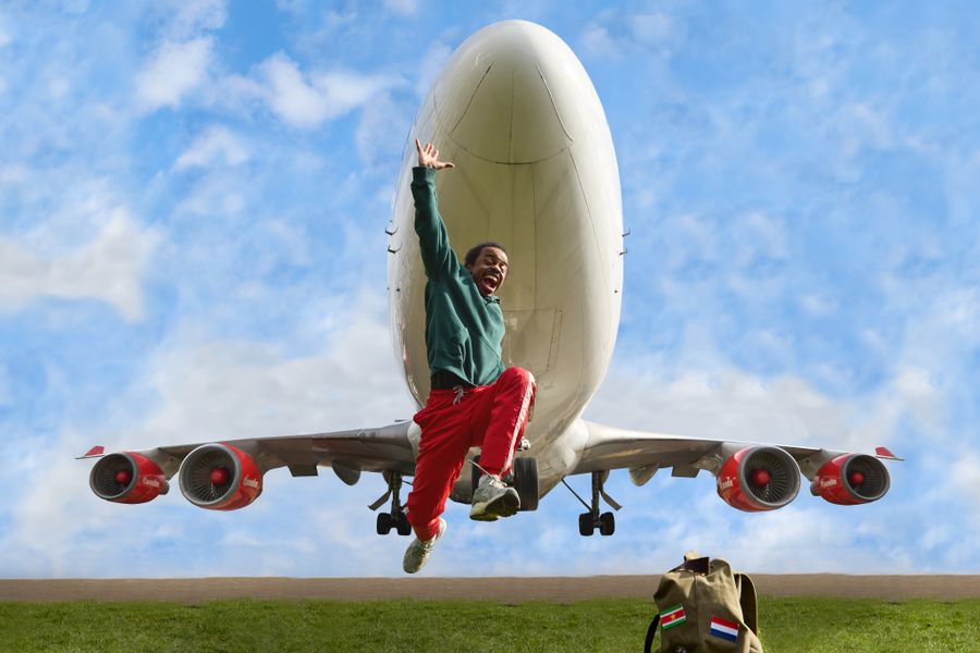 Een jonge man met een extatisch blije blik op zijn gezicht springt in de lucht met 1 vuist omhoog vlak voor  de neus van een vliegtuig dat lijkt te zweven vlak boven de grond. de grond zelf is een groene grasmat met een ongedefinieerde bruine strook aan de horizon en daarboven een blauwe lucht met witte wolkjes. Voor de man staat een grote rugtas met vier badges, waaronder een smiley en een Nederlandse en Surinaamse vlag.