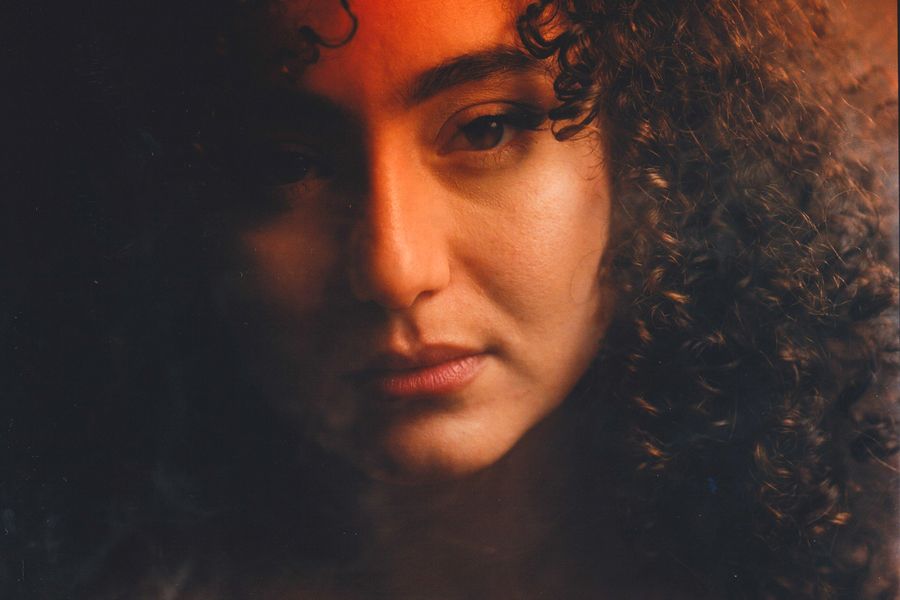 Een vrouw met donker krullend haar kijkt in de camera en wordt met een warme oranje-achtige gloed aangelicht