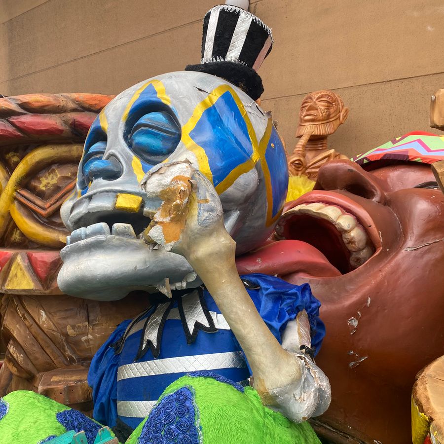 Grote carnavalsfiguur van een blauw-grijze schedel met felgekleurde details, zittend tussen andere beschadigde decorstukken.