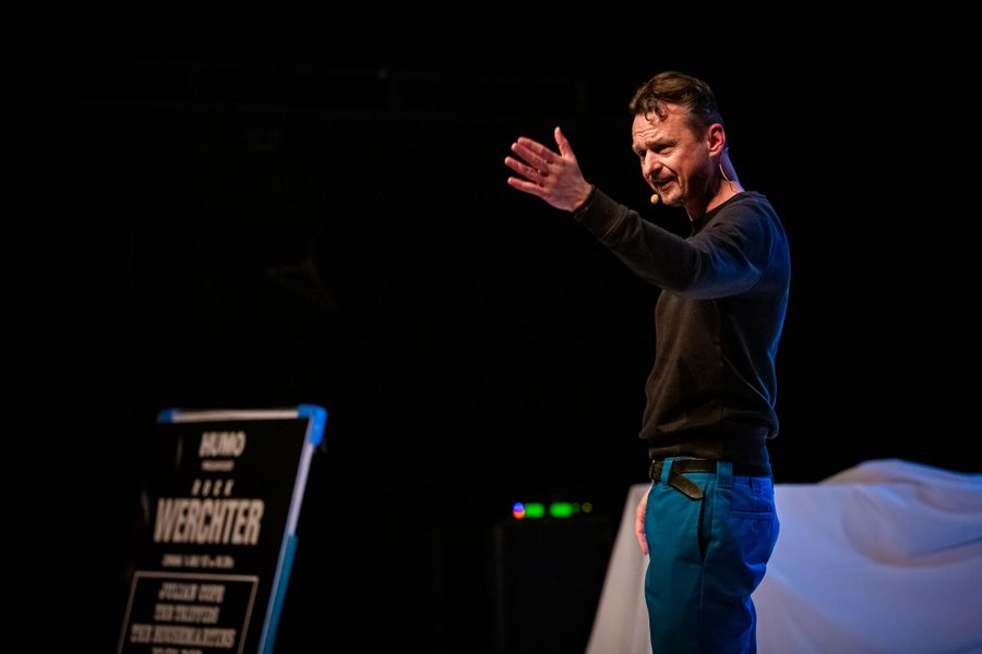 De Cock staat op een podium met een microfoonheadset en maakt een expressief gebaar met zijn hand, alsof hij het publiek toespreekt of een punt kracht bijzet. Op de achtergrond is een bord zichtbaar met "Rock Werchter".