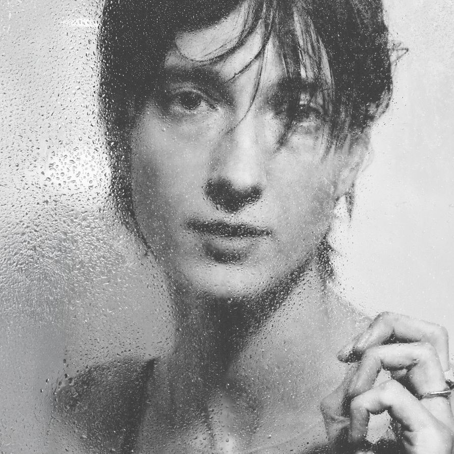 Zwart-wit portret van een persoon achter een beslagen en nat oppervlak, met waterdruppels op het glas en een dromerige, zachte blik.