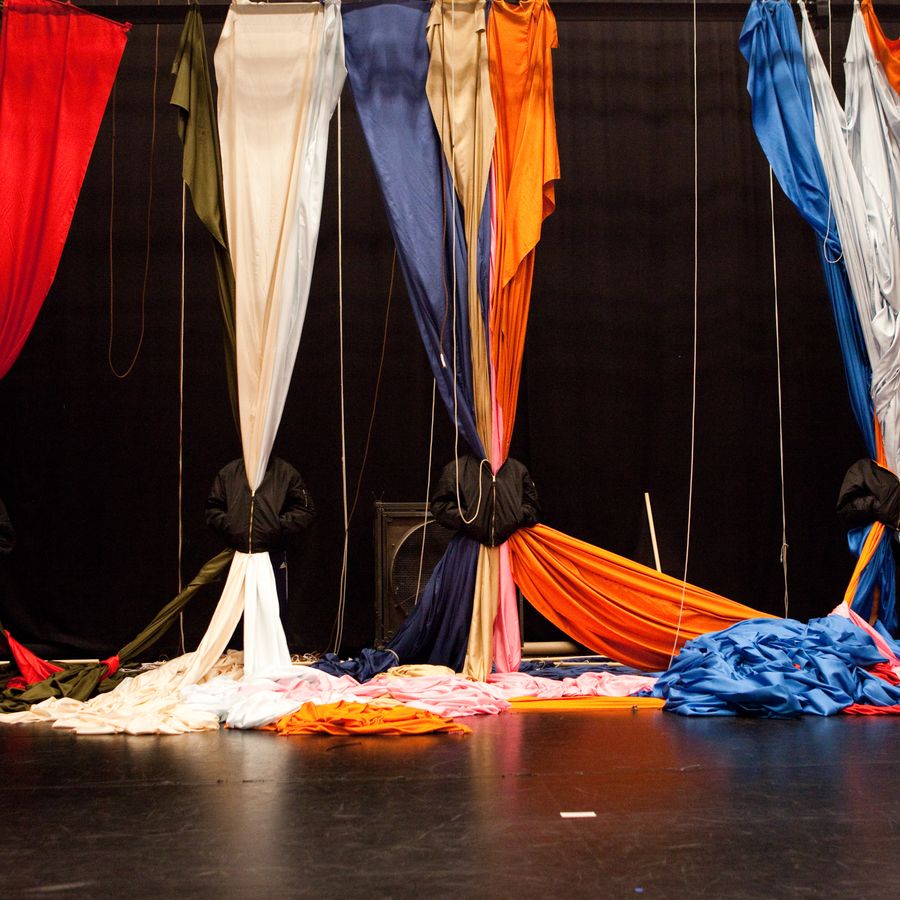 Vier mensen in zwarte bomberjacks tegen een zwarte achtergond - en daardoor bijna onzichtbaar - hebben grote hangende doeken tussen hun jas hangen waardoor behalve de jassen de personen geheel aan het zicht worden onttrokken