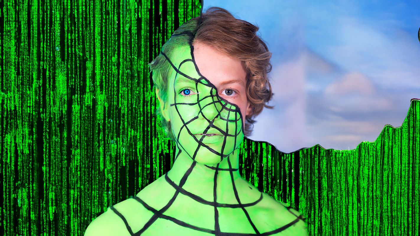 Een jong mens met krullend haar kijkt in de camera, het grootste deel van diens gezicht en schouders wordt bedekt door een groen spidermanpak, daarachter is een zwartgroene achtergrond van computercode te zien die kenmerkend is voor The Matrix. Achter het onbedekte deel van zijn gezicht is een blauwe wolkenlucht te zien.